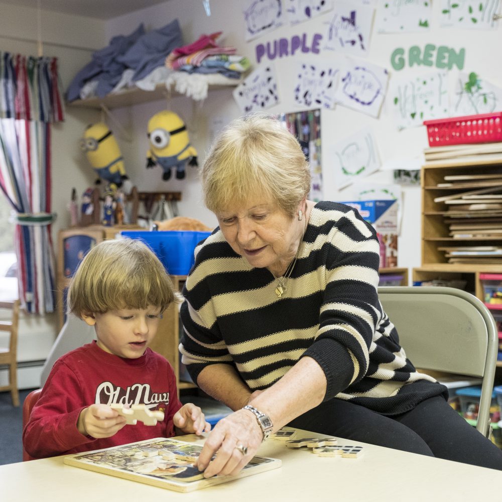 An older woman helps a young child finish a puzzle in a classroom