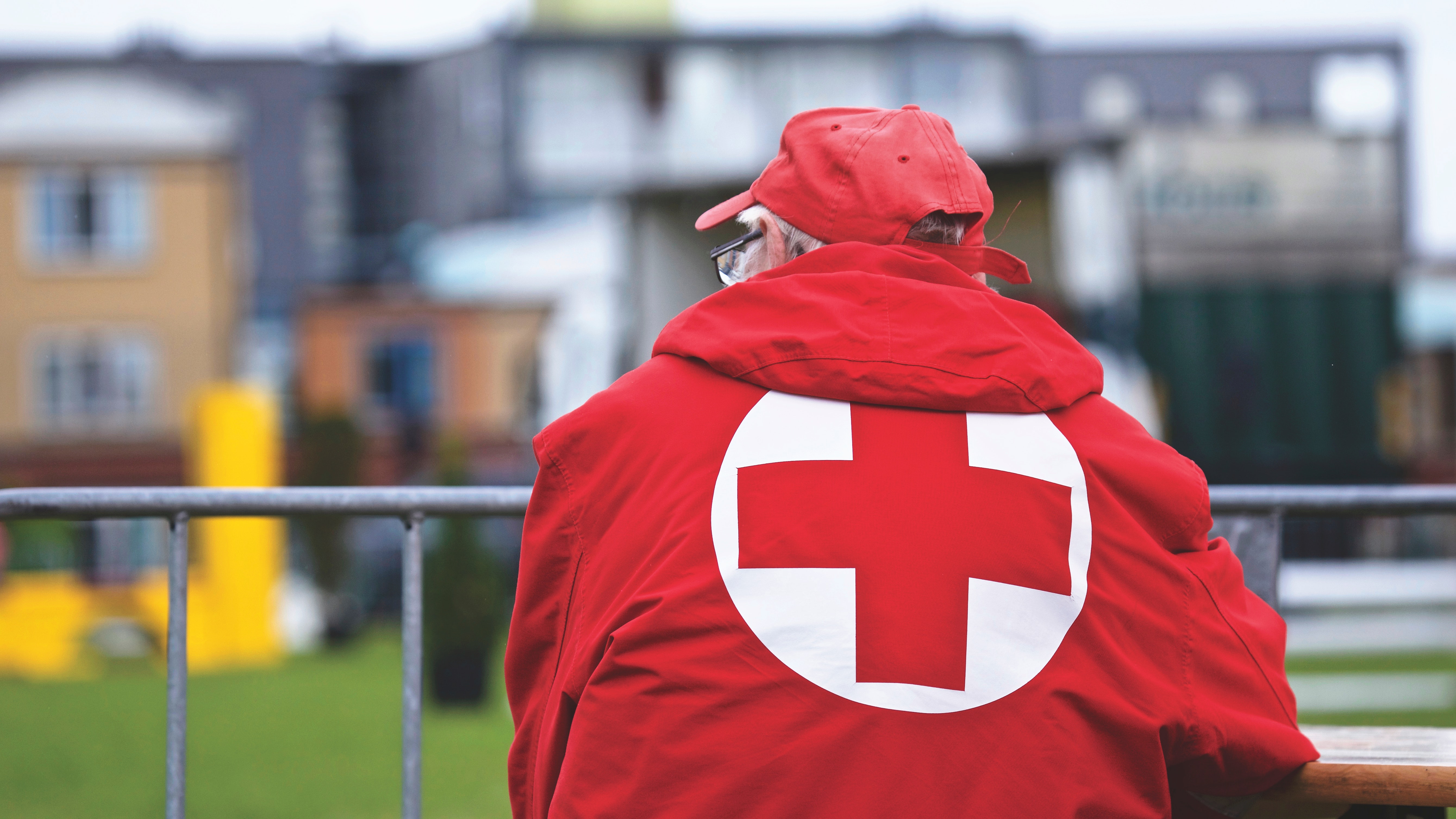 An older man in a red jacket with the Red Cross logo on his back looks out onto a field